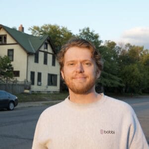This is a headshot of me from the chest up, after a nice haircut, at sunset on my street in Northeast minneapolis. There are a few cars and church steeple, as well as trees, lining the background.